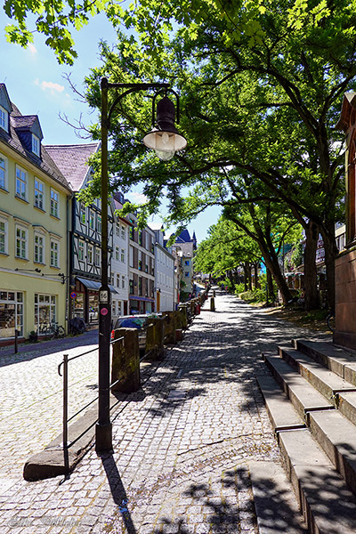  Eine leicht ansteigende, sonnenbeschienene Kopfsteinpflasterstraße in Marburg, gesäumt von historischen Gebäuden auf der linken Seite und hohen grünen Bäumen auf der rechten Seite. Eine alte Laterne steht im Vordergrund, der Blick reicht eine Gasse hinauf.