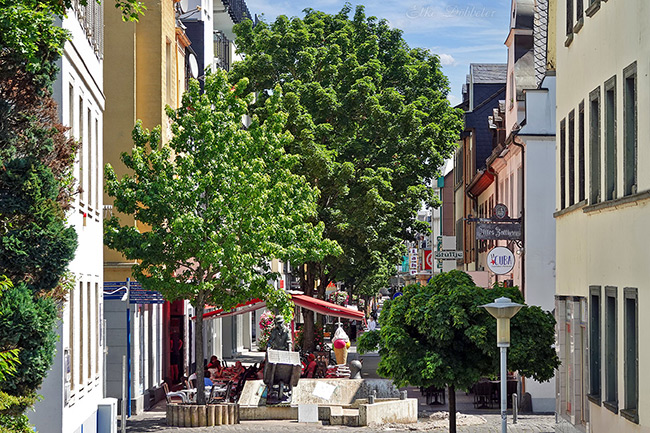Sonnige Straßenszene in Neuwied mit dem Scherjer-Brunnen im Vordergrund und einem belebten Straßencafé unter Bäumen.