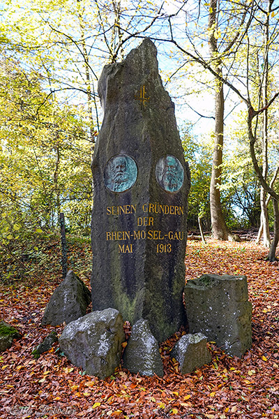 Detaillierte Aufnahme des moosbewachsenen Denkmals auf dem Krahnenberg mit der Inschrift: "Seinen Gründern der Rhein-Mosel-Gau Mai 1913". Zwei metallene Porträt-Medaillons sind eingelassen.