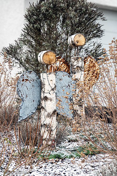 Mehrere schlichte Engelsfiguren aus Holz und Metall stehen im Freien im Schnee, dezent dekoriert und ruhig in die winterliche Umgebung eingebettet.