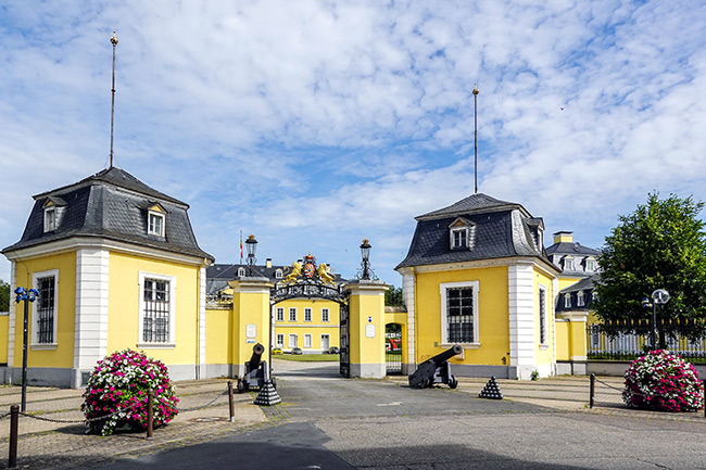 Historische Ansicht des Schlosses Neuwied bei blauem Himmel. Vor den gelben Torhäusern stehen zwei alte Kanonen und große, blühende Blumenkübel. Das Pflaster wirkt unebener und lückenhafter als auf dem aktuellen Foto.