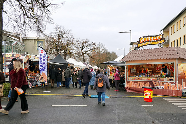 Blick über das Festivalgelände in einer Stadtstraße. Zwischen den Ständen, darunter ein großer Wagen für spanische Churros, schlendern viele Besucher unter bewölktem Himmel. Foto: Elke Döbbeler