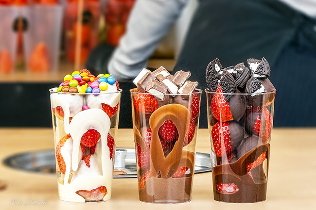 Drei Klarsichtbecher gefüllt mit frischen Erdbeeren, die großzügig mit flüssiger weißer, Vollmilch- und Zartbitterschokolade überzogen und mit bunten Schokolinsen oder Keksen getoppt sind. Foto: Elke Döbbeler