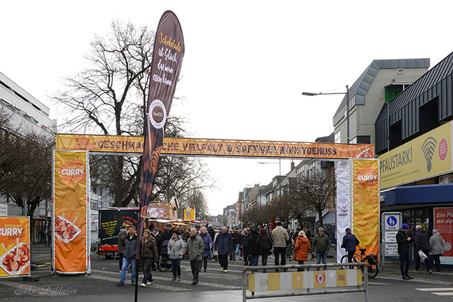 Ein großer, orangefarbener Torbogen markiert den Eingang zum Currywurstfestival in einer belebten Fußgängerzone. Menschen schlendern bei bewölktem Wetter zwischen den Ständen. Foto: Elke Döbbeler