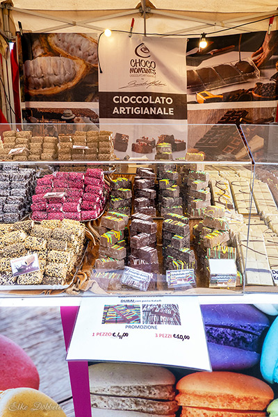 Eine bunte Auswahl an handgemachter Schokolade (Cioccolato Artigianale) hinter einer Glasscheibe. Zu sehen sind verschiedene Riegel mit Toppings und Dubai-Schokolade. Foto: Elke Döbbeler