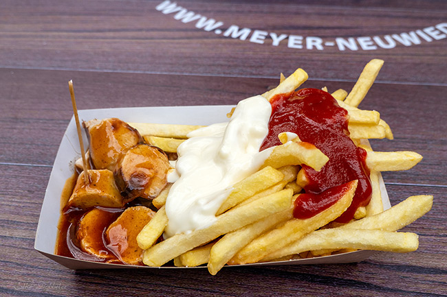 Eine Portion Currywurst mit Pommes rot-weiß in einer weißen Schale. Die Pommes sind großzügig mit Ketchup und Mayonnaise bedeckt. Foto: Elke Döbbeler
