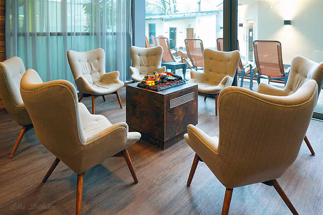 Gemütlicher Ruheraum mit einem modernen Elektrokamin in der Mitte, umgeben von hellen Polstersesseln auf Holzboden.