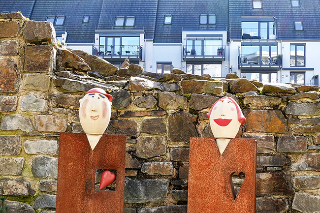 Zwei abstrakte Skulpturen aus rostigem Metall mit Köpfen aus heller Keramik stehen vor einer alten Bruchsteinmauer. Die linke Figur trägt eine rote Kappe und hat ein kleines rotes Herz in einer quadratischen Aussparung im Brustbereich; die rechte Figur hat rote Haare und eine herzförmige Aussparung. Beide lächeln freundlich und blicken direkt in die Kamera. Im Hintergrund ist eine moderne, hellgraue Wohnanlage mit Balkonen und dunklem Dach zu sehen, die leicht dunstig wirkt. Ein charmantes Detail: Am Kopf der linken Figur ist eine feine Spinnwebe zu erkennen.